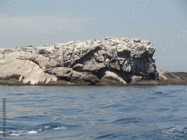 Fototapeta Large gray and white fractured rock in the middle of the ocean. Blue sea, blue sky. Laje de Santos, the whale-shaped rock at Brazil, also a diving venue. Human face profile shape on the right. 