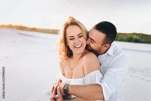 Fototapeta Brodaty mężczyzna i blond kobieta przytulają się na tle białego piasku. Portret emocjonalnych ludzi o zachodzie słońca. Miłość na pustyni nowożeńców. Historia miłosna wesołych i młodych kochanków.