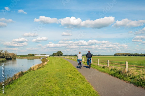 Fototapeta Two unidentified people cycle on a bike path at the top of a dike