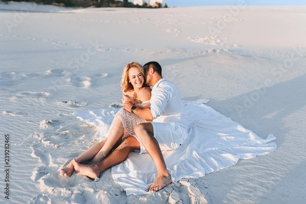 Obraz A bearded man and a blond woman hug sitting on the white sand and enjoy the sunset. Love in the desert newlyweds. The love story of fun and love people.