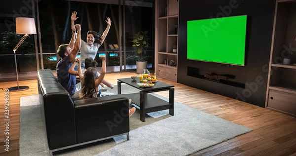 Obraz A family is watching a TV and celebrating some joyful moment, sitting on the couch in the living room.