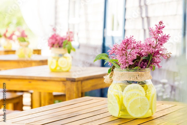 Fototapeta Bouquet of lilac flowers in a jar on the table. Jar filled with water with lemon slices. Photo in bright colors. Decoration for restaurants.
