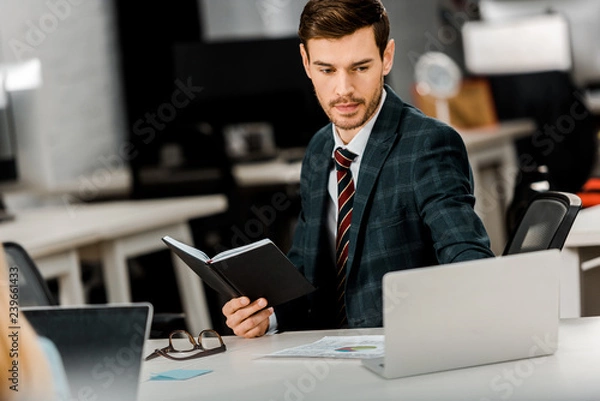 Fototapeta focused businessman with notebook working on laptop in office