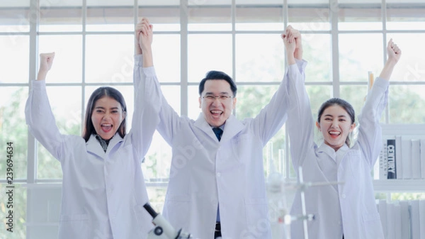 Obraz The powerful scientists hold hands raised in colleague lab. They are smiling congratulation for success experiment in biotechnology and pharmacy in career. Science, chemist, biology education concept.