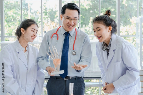 Obraz A handsome professor doctor teaching couple of beautiful young asian girl collage student in white laboratory gown learning anatomy class.  Anatomic bone of  hand in biology classroom in university.