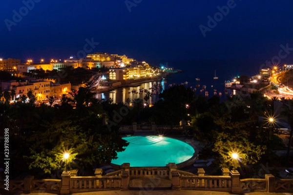 Fototapeta View of St. George's Bay seafront lights by night, with a blue luxury swimming pool and boats and yachts anchored. St Julian's (San Giljan), Central Region, Malta. Evening in Paceville district