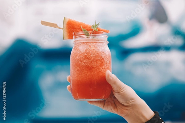 Fototapeta Hand with Fresh watermelon smoothie in the glass jar. Background is blur swimming pool