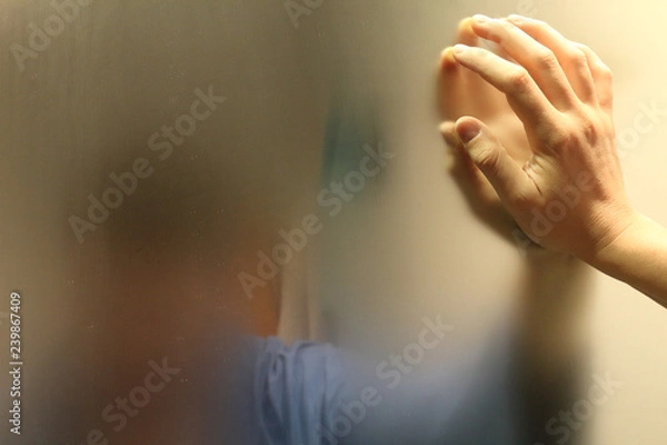 Fototapeta A meditative man with a blue bathrobe in a bathroom watching himself in a foggy blurry mirror and resting his hand on it, in an introspective moment