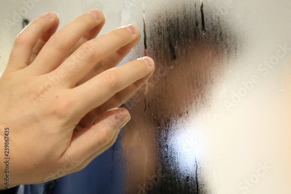 Fototapeta A meditative man with a blue bathrobe in a bathroom watching himself in a foggy blurry mirror and resting his hand on it, in an introspective moment