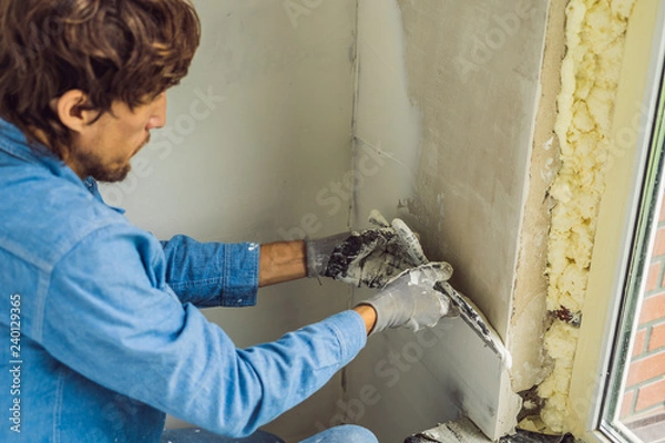 Fototapeta master is applying white putty on a wall and smearing by putty knife in a room of renovating house in daytime