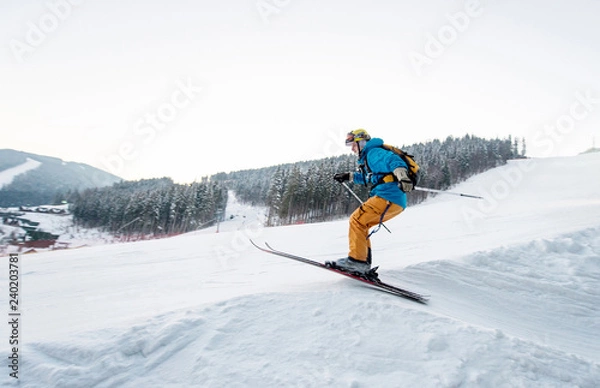 Fototapeta Skier man at jump from the slope of mountains in blue jacket with forest in background. Side view