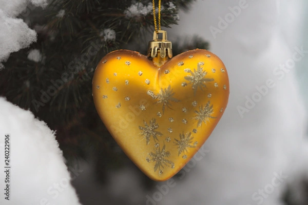 Fototapeta Christmas decoration in the form of a golden heart with patterns on the background of snow-covered coniferous tree close-up