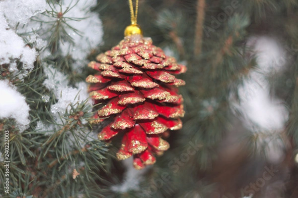 Fototapeta red Christmas pine cone with gold spangles on the background of snow-covered coniferous tree close-up