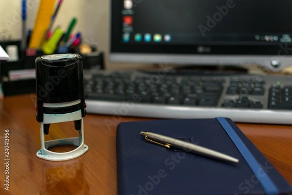 Fototapeta Stamp for documents, notepad and pen lie on a wooden office desk on a computer and stationery background, closeup