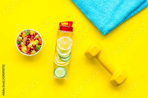 Obraz Healthy lifestyle, healthy habits. Detox water, fruit salad, sport equipment dumbbells on yellow background top view