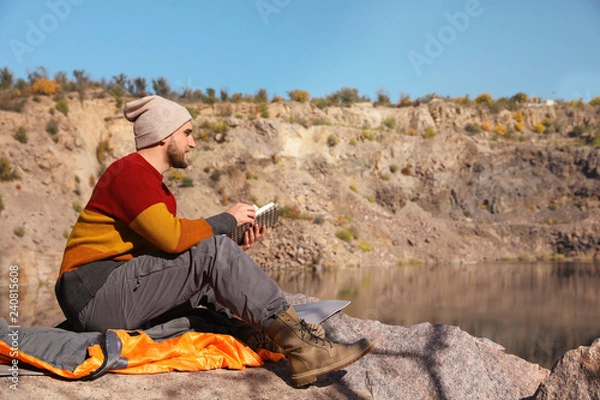 Fototapeta Male camper reading book while sitting on sleeping bag in wilderness. Space for text