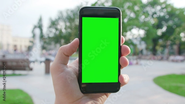 Fototapeta Lublin, Poland - July 2018: A hand holding a phone with a green screen in a public park. Close-up shot. Soft focus