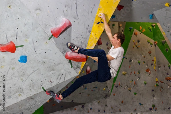 Fototapeta Rock climber man hanging on a bouldering climbing wall, inside on colored hooks