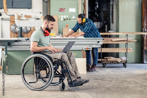 Obraz worker in wheelchair with computer in a carpenter's workshop with his colleagu
