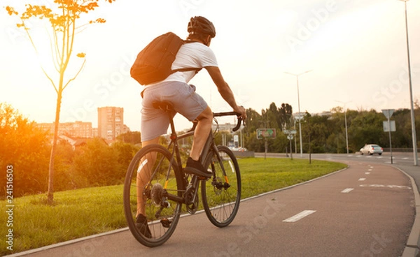 Fototapeta The young guy in casual clothes is cycling on the road in the evening city