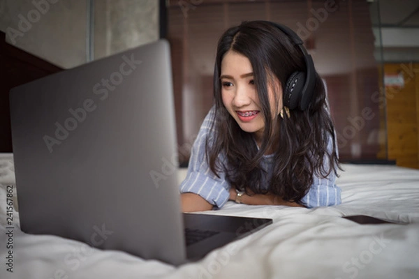 Fototapeta Girl with headphones listening music or watching movie with laptop on the bed at home.Young beautiful asian woman lying down relaxing and smiling with notebook computer and headset in luxury room.