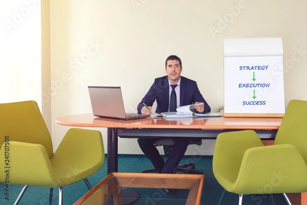 Fototapeta Confident young team leader or manager sitting at desk next to laptop and flip chart smiling while working with documents and taking notes – concept of business strategy, success – warm filter