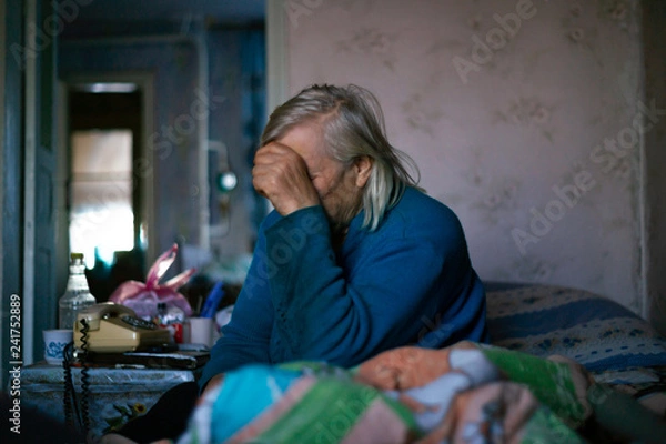 Fototapeta Old poor gray hair woman covered her face with her hand. Woman is sad. Poor life in village. Old age not good. Low-light photo.