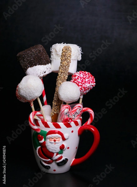 Obraz White Christmas mug with a Santa Claus picture filled with cake pops, marshmallows and candy canes on a black background with text space. Christmas baked gifts.