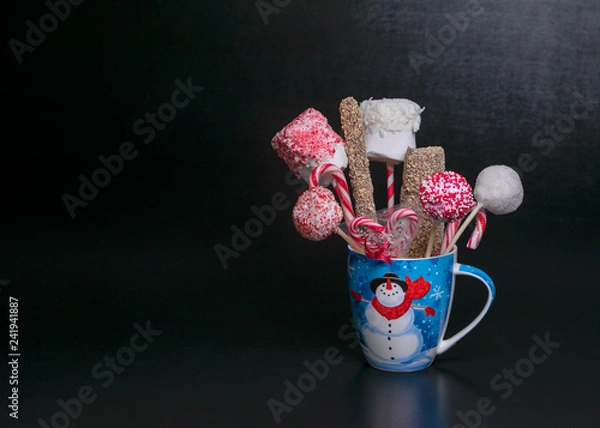 Obraz Blue Christmas mug with a snowman filled with candy pops, marshmellows and candy canes on a black background with text space.  Christmas baked gifts.