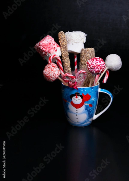 Obraz Blue Christmas mug with a snowman filled with candy pops, marshmellows and candy canes on a black background with text space.  Christmas baked gifts.