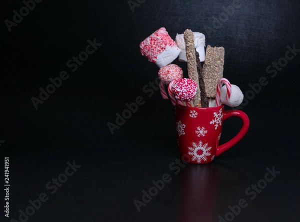 Obraz Red Christmas mug with snowflakes filled with cake pops, candy canes and marshmallows on a black background with text space. Christmas baked gifts.