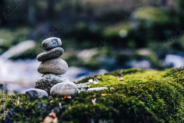 Fototapeta Balancing rock art. Stones balanced on top of each other on the stone with moss. Green color, flowing creek and a stone man. Zen stones or zen stack in the forest next to a river.