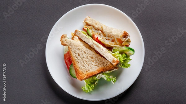 Obraz Homemade sandwich made of toast bread, fried egg, melted cheese, tomatoes, cucumber, lettuce, burgers and mayonnaise sauce. Sandwich on a white plate, on a black background. top view