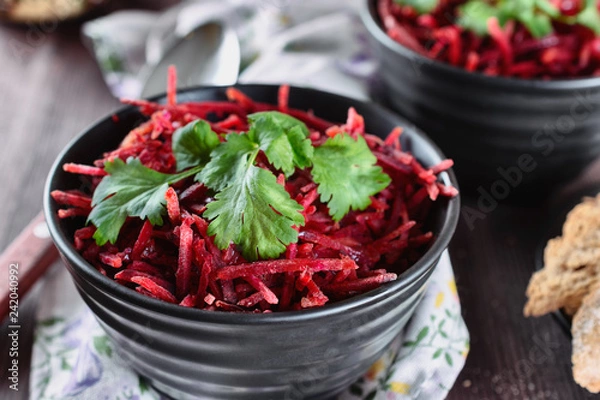 Fototapeta Thinly sliced ​​fresh beets in a black salad bowl with sprigs of cilantro ready for eating are on a wooden table. Shredded beet salad, closeup.