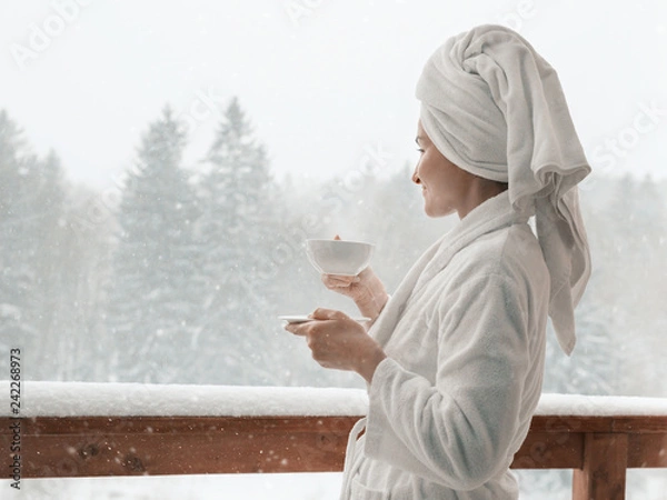 Fototapeta Young woman on the balcony holding a cup of coffee in the morning. She in hotel room looking at the nature in winter. She is with the bath towel on her head an wearing bath coat.