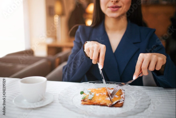 Fototapeta Cut view of nice young woman cut some cake with knife and fork. She sit at table in restaurant. Cup of coffee stand there. She enjoy her meal.