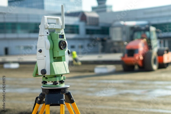 Fototapeta Surveyor equipment (theodolite) on construction site of the airport, building or road with construction machines in background