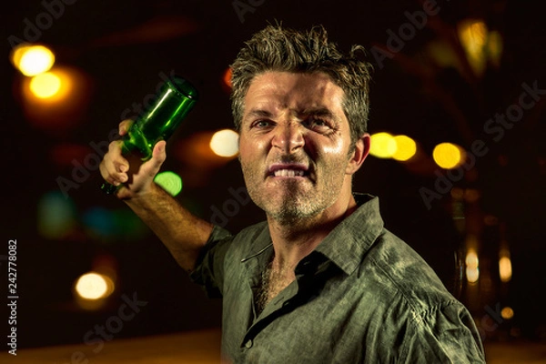 Fototapeta young upset and aggressive drunk man in pub at night holding beer bottle threatening ready to fight as the violent bar thug troublemaker and alcohol addict furious guy