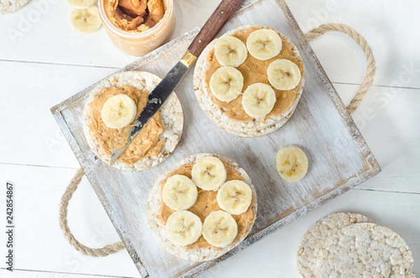 Obraz Healthy breakfast with rice cakes with peanut butter and slices of banana on white wooden table. Top view.