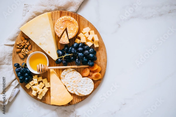 Fototapeta Flat lay with various types of cheese, grapes, nuts, honey and cracker in wooden board on marble