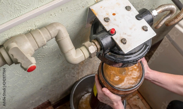 Fototapeta Plumber unscrews glass flask with old water filter in basement.