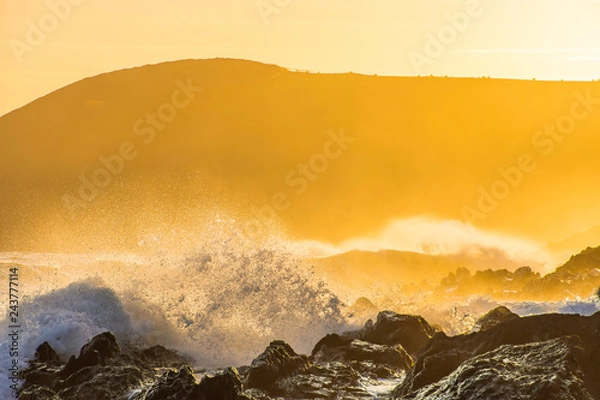 Obraz Sunset on Pembrokeshire coast.Big waves splashing on rocky shore and mist over sea with cliff in background during golden hour.