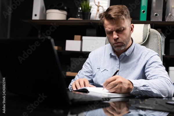 Fototapeta young man sitting at computer table and makes notes in diary