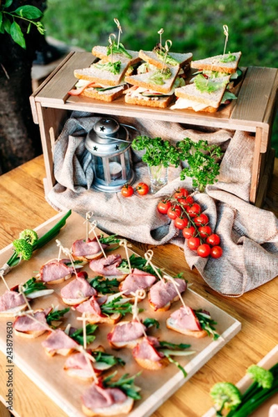 Fototapeta Delicious catering banquet buffet table decorated in rustic style in the garden. Different snacks, sandwiches with ham, chiken, cheese and greenery on a wooden plate. Outdoor. Vertical photo.