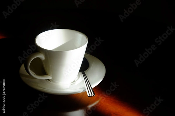 Fototapeta Empty white coffee glass with saucer and metal spoon in the dark corner with beautiful sunlight on wooden glossy table