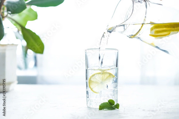 Obraz Pouring a glass of water with lemon, ice and mint on a white table