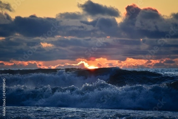 Fototapeta Storm on the sea at sunset. Blue sky with dark clouds. The light of the sun on the horizon. Big waves roll ashore. Splashes and mist over the wave.
