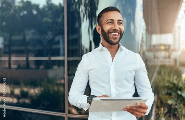 Fototapeta Portrait of a young confident smiling indian man holding a tablet and looking into the distance