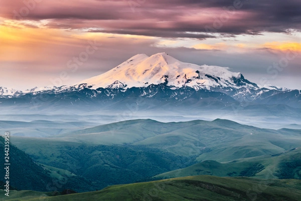 Fototapeta Beautiful view of Elbrus mount in the morning at sunrise with a dramatic cloudy sky and foggy green highland meadows. Summer mountain landscape. North Caucasus, Karachay-Cherkess Republic, Russia
