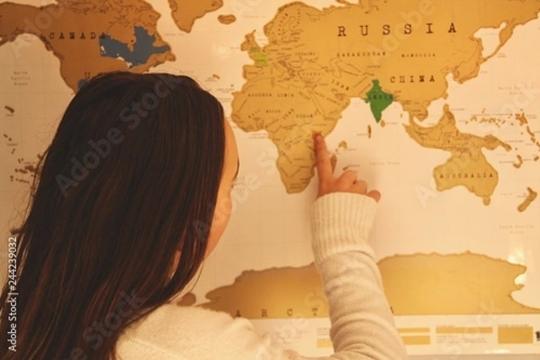 Fototapeta Young teenage girl pointing at a destination on a scratch map of the world on the wall